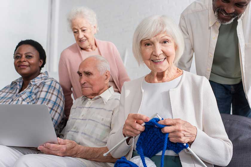 Elderly woman holding yarn near multiethnic friends with laptop in nursing home